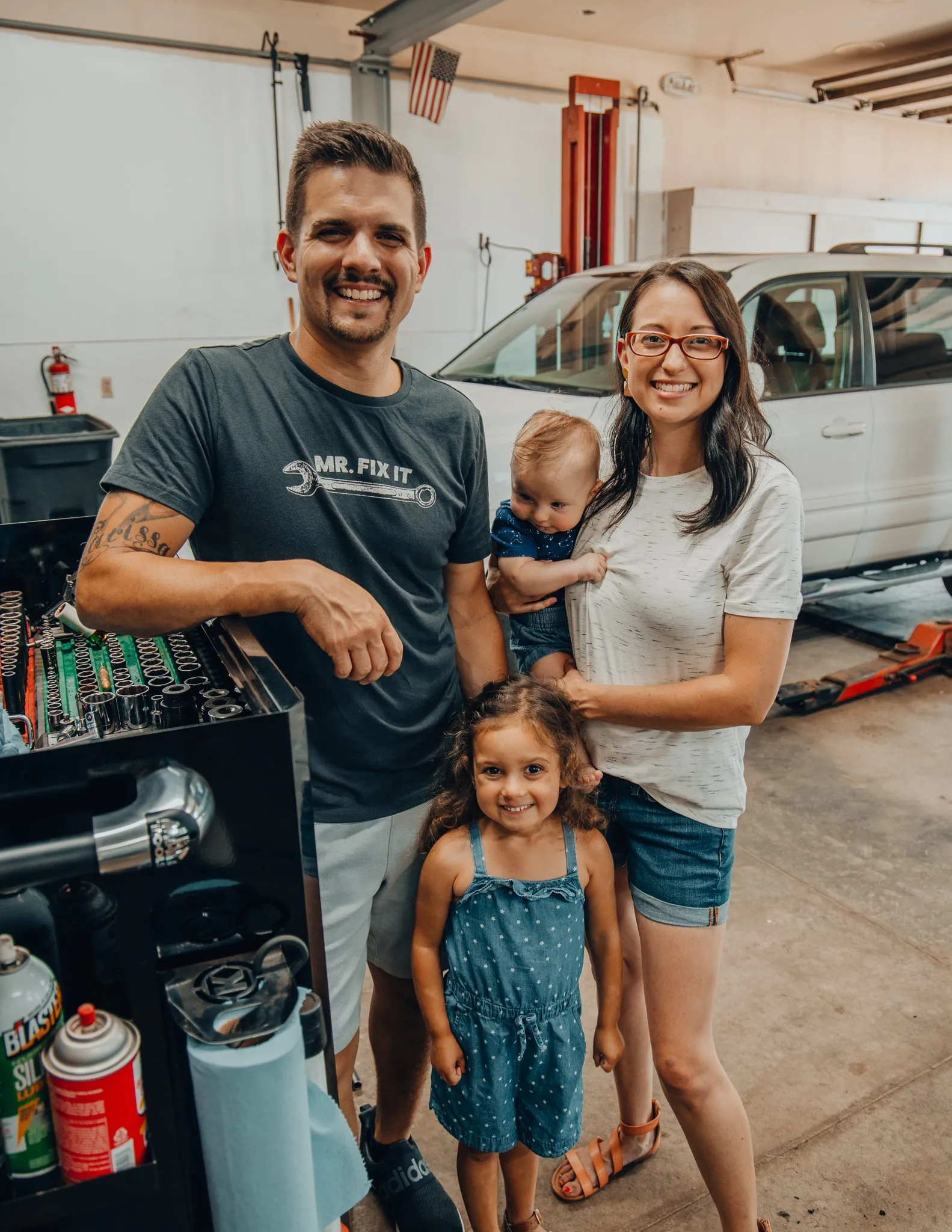 Enright Automotive shop owner Chris Enright with wife and kids. Also in the image are shop tools and a white car in the background.