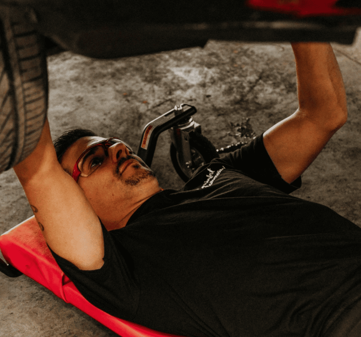 Closeup image of shop owner Chris Enright on a rolling bed under a lifted car that's under repair. Concept image of Honda and Acura repair and preventative maintenance at Enright Automotive.
